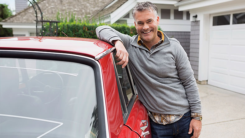 Grey haired man leaning against the window of a red car parked in suburban driveway