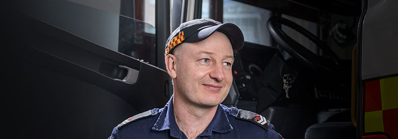 An emergency services worker standing in front of his vehicle with a small smile looking off camera.