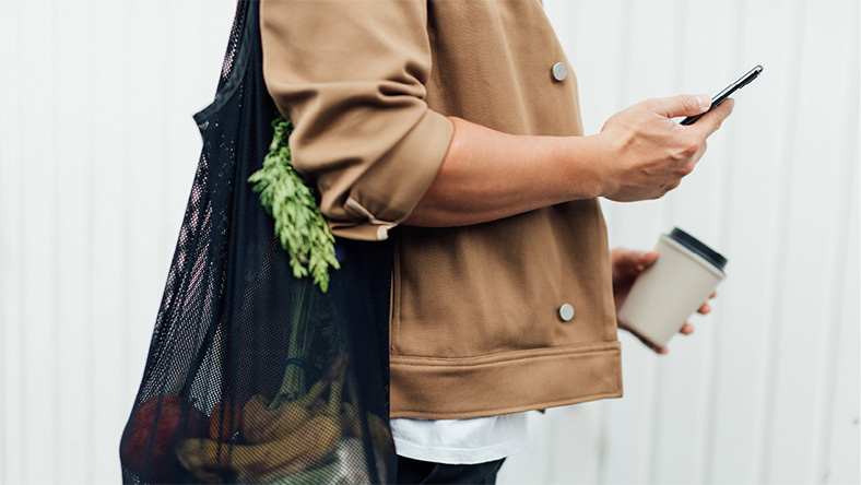 A side shot of a person on their phone while practicing sustainable lifestyle habits by using a reusable shopping bag and coffee cup.