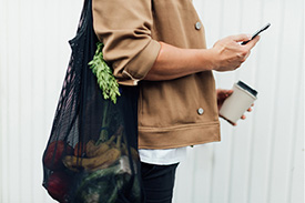 A side shot of a person on their phone while practicing sustainable lifestyle habits by using a reusable shopping bag and coffee cup.