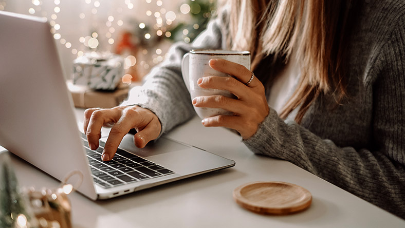 A young woman shops safely on her laptop while clutching a mug of coffee