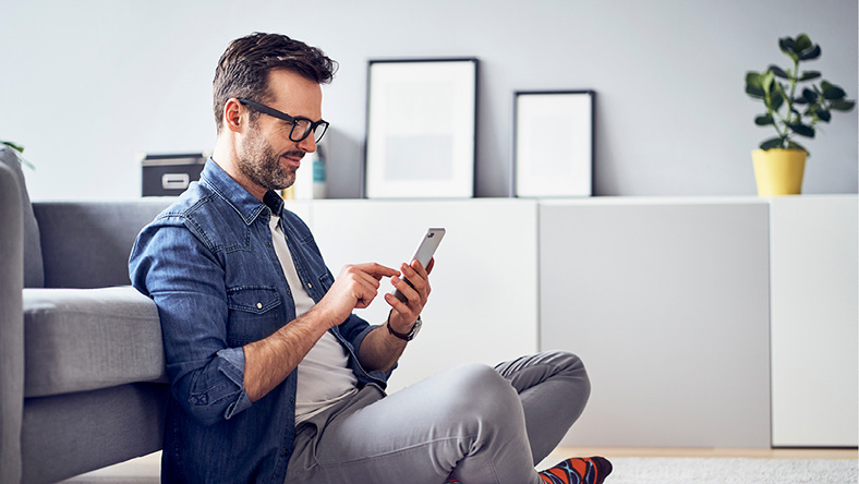 A man sits on the floor f his living room using personal finance banking apps on his phone.