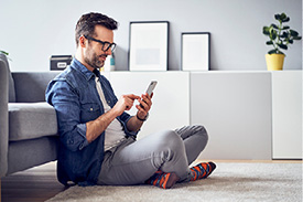 A man sits on the floor f his living room using personal finance banking apps on his phone.