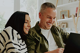 A couple using a laptop sitting on a lounge smiling.