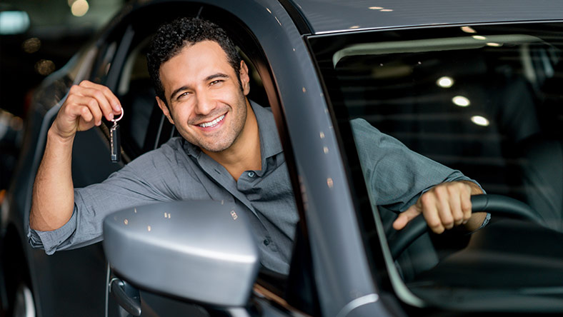 A man with dark hair has his arm out of a new car window holding his car keys.