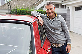 Grey haired man leaning against the window of a red car parked in suburban driveway