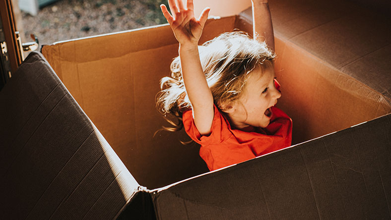 A happy child raises her hands while sitting in a cardboard box.
