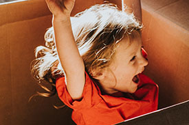 A happy child raises her hands while sitting in a cardboard box.