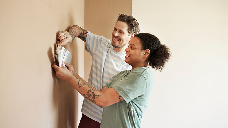 A young couple hold hold a paint swatch chat against a wall in their home.