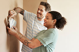 A young couple hold hold a paint swatch chat against a wall in their home.