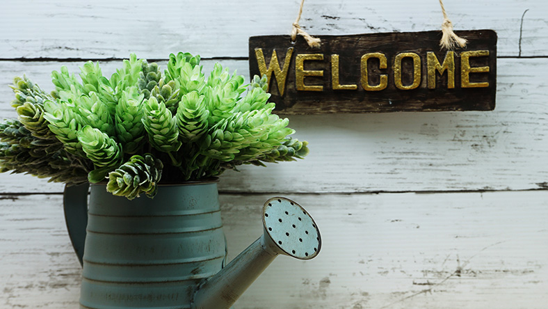 A watering can with a succulent flowering from the top sits beneath a welcome sign