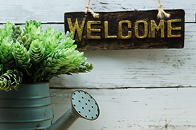 A watering can with a succulent flowering from the top sits beneath a welcome sign