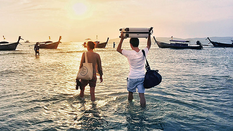 A man and woman knee deep in water, walking out to boat with their luggage in tow