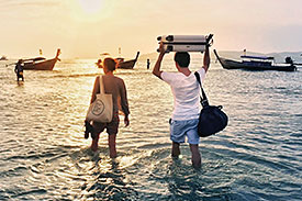 A man and woman knee deep in water, walking out to boat with their luggage in tow