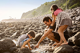 Father plays with two children on a pebbly beach