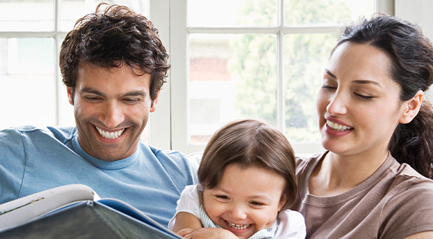 A man in a blue t shirt and his partner, a woman in a brown shirt, sit on a couch with their baby girl. All three are smiling and reading a book.
