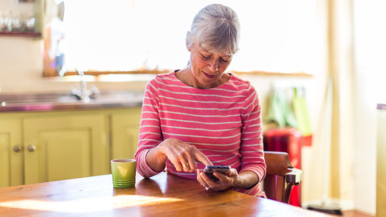 A woman sits at her kitchen table using her mobile phone.