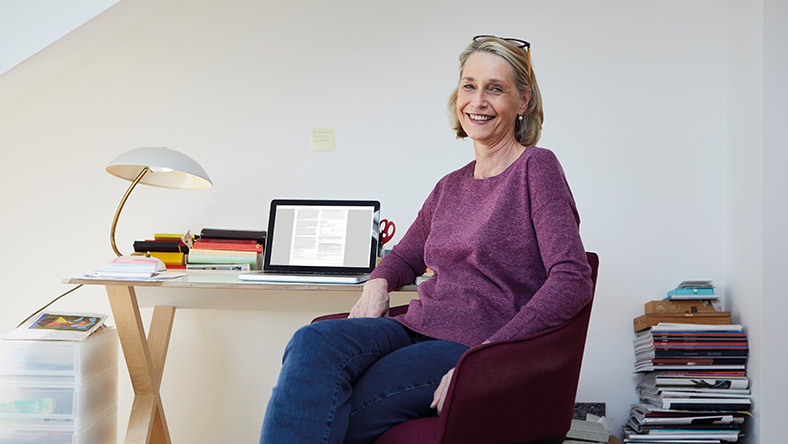 A woman sits at her desk with her laptop and smiles at the camera.