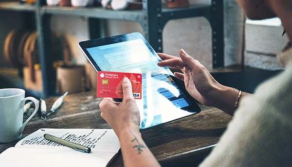 A close up of someone using an iPad and holding a red Firefighters Mutual Bank Credit card. In the background is a mug, clay sculpting tools and a wooden bench.