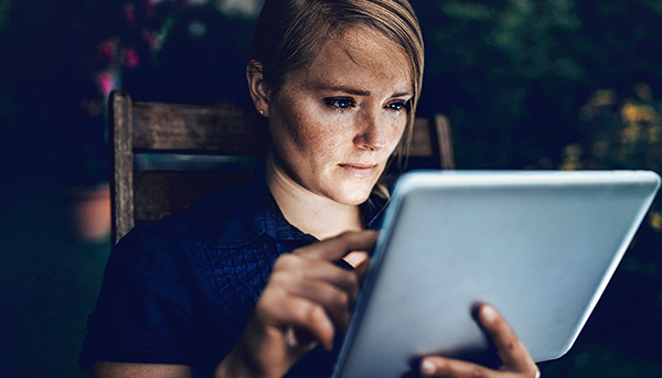 A close up image of a serious-looking woman using a tablet device while sitting in a chair. The background is dark.