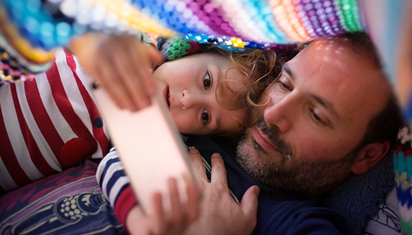 A close up image of a father and child looking at a mobile phone. A colourful blanket is draped over them.