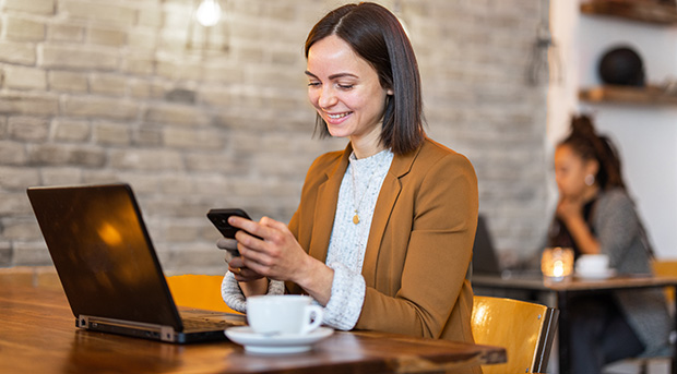 A woman works on a laptop in a cafe while securely checking her accounts in the Firefighters Mutual Bank app.