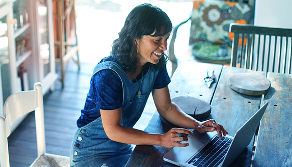 A woman wearing overalls and a blue t-shirt sits at a wooden table using a laptop and smiling. Shelves and an armchair are visible in the background. 