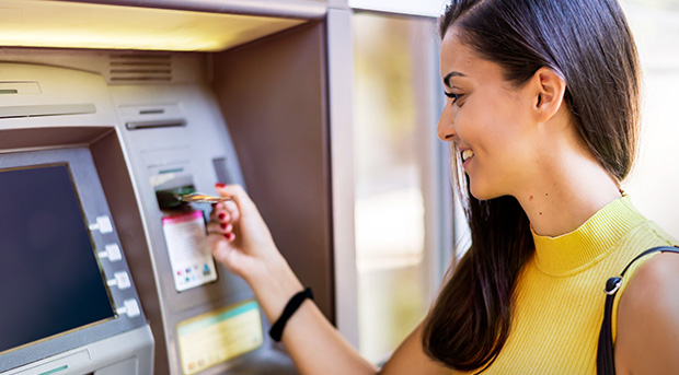 A young woman in a yellow top inserts her bank card into an ATM.
