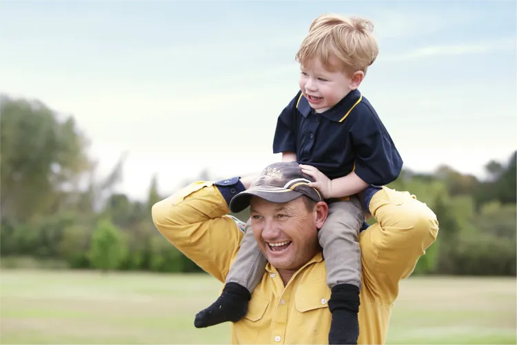 A man in a yellow shirt carries a young boy on his shoulders in an outdoor setting.