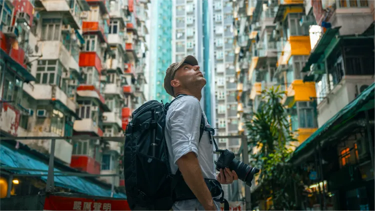 An adult male stands in a densely packed urban environment, holding a camera and wearing a large backpack.
