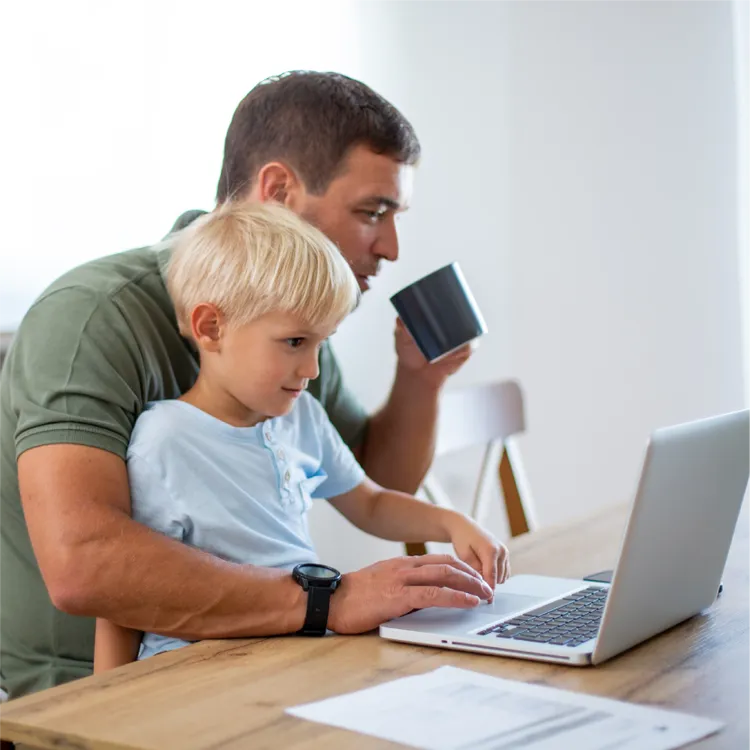 An adult sits at a wooden table with a child on their lap, both focused on a laptop.