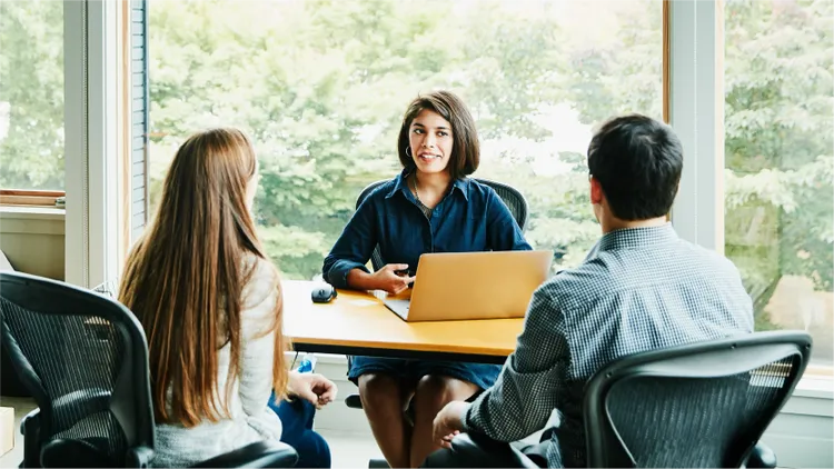 Three colleagues sit together around a table in a bright office space.
