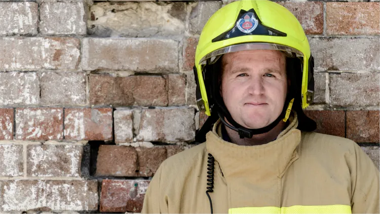 A firefighter wearing a bright yellow helmet and tan protective suit is positioned against a weathered brick wall.