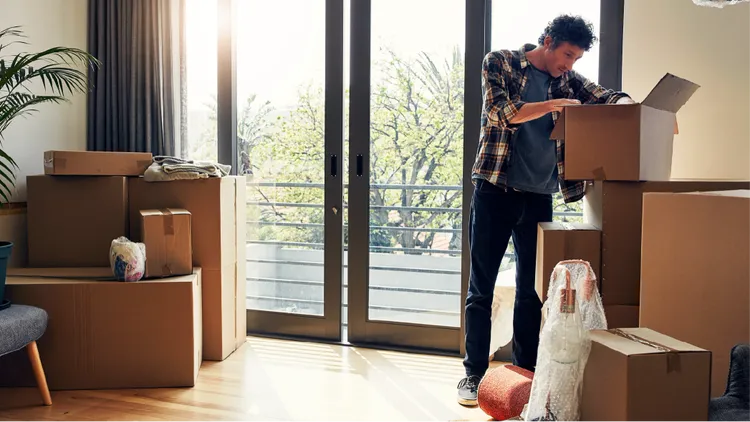 A young adult man stands in a bright apartment opening a cardboard box.