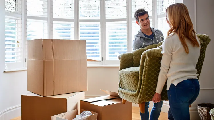 Two adults carry a green upholstered armchair together in a bright room.