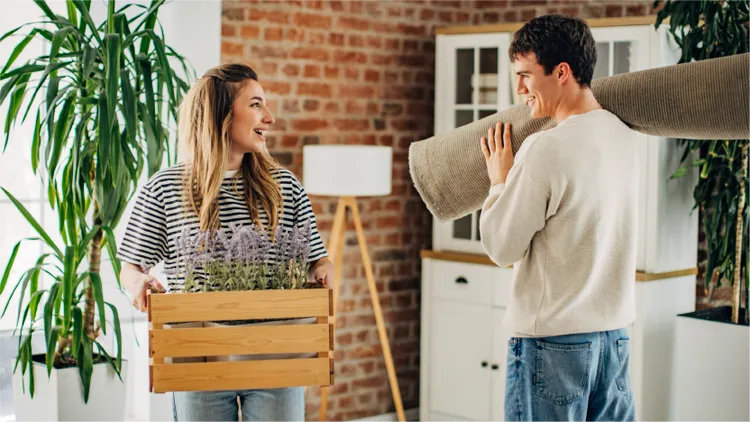 Two adults move into an apartment, carrying a wooden crate of potted plants and a rolled-up rug near an exposed brick wall.