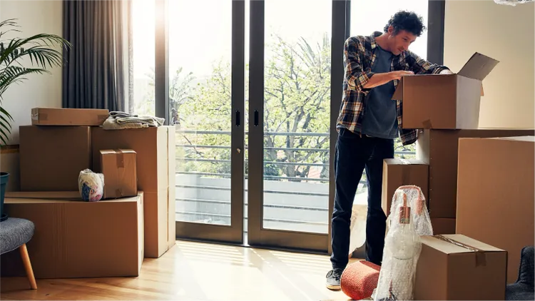 A young adult man stands in a bright apartment opening a cardboard box.