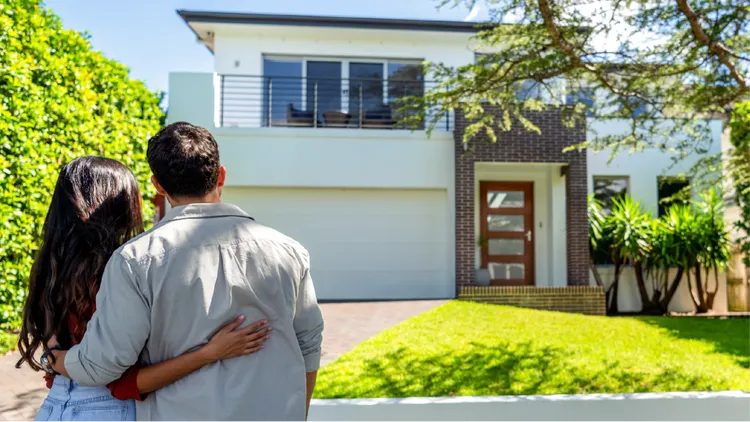 A young couple stands arm in arm, looking at a modern two-story house.