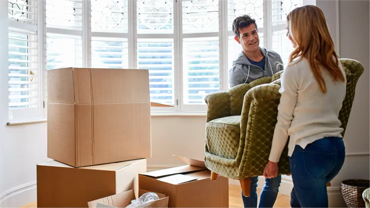 Two adults carry a green upholstered armchair together in a bright room.