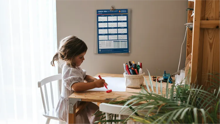 A young child sits at a small wooden desk, drawing on a blank sheet of paper.