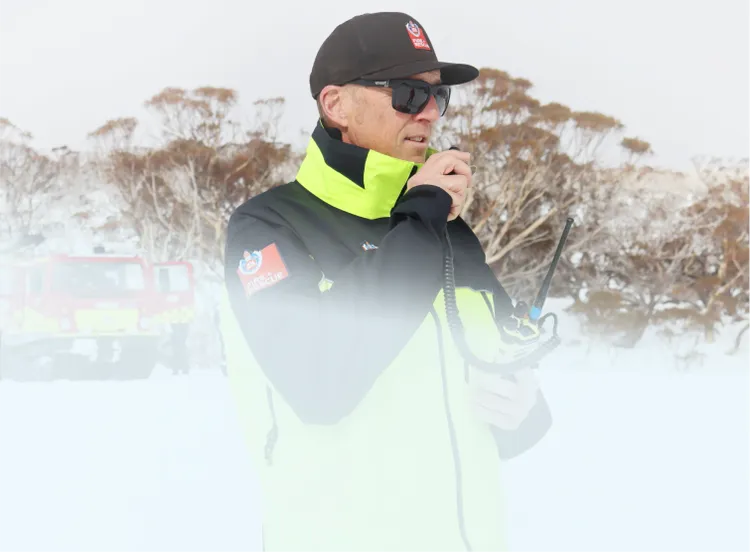 A rescue worker in a bright high-visibility jacket stands in a snowy landscape using a handheld radio.