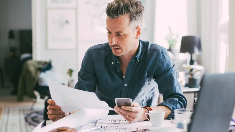 A casually dressed man sits at a table reviewing printed documents while holding a smartphone.