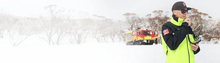 A rescue worker in a bright high-visibility jacket stands in a snowy landscape using a handheld radio.