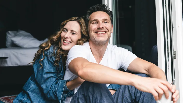 A young couple sits closely together near an open doorway, with natural light illuminating them from outside.