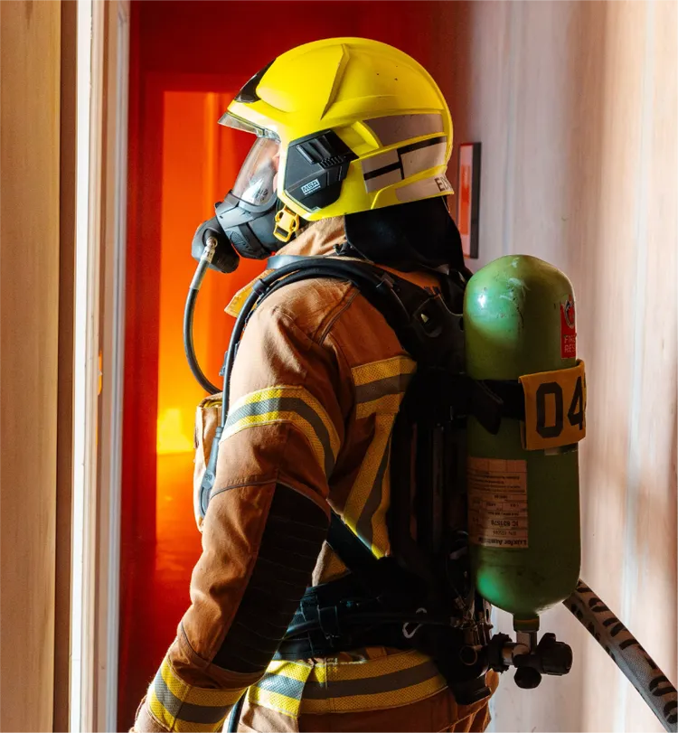 A firefighter in full protective gear stands in a narrow hallway, facing a brightly lit doorway.