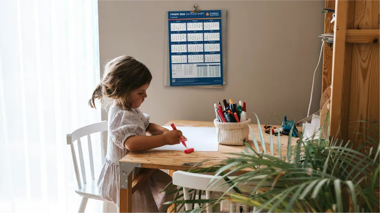 A young child sits at a wooden table, focused on drawing with a red marker.