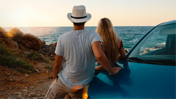 A young couple stands beside a car, looking out over the ocean at sunset.