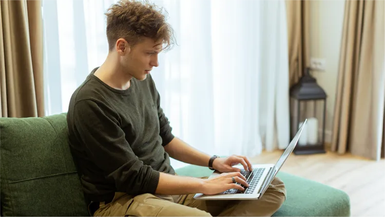 A young man sits on a green sofa using a laptop computer.